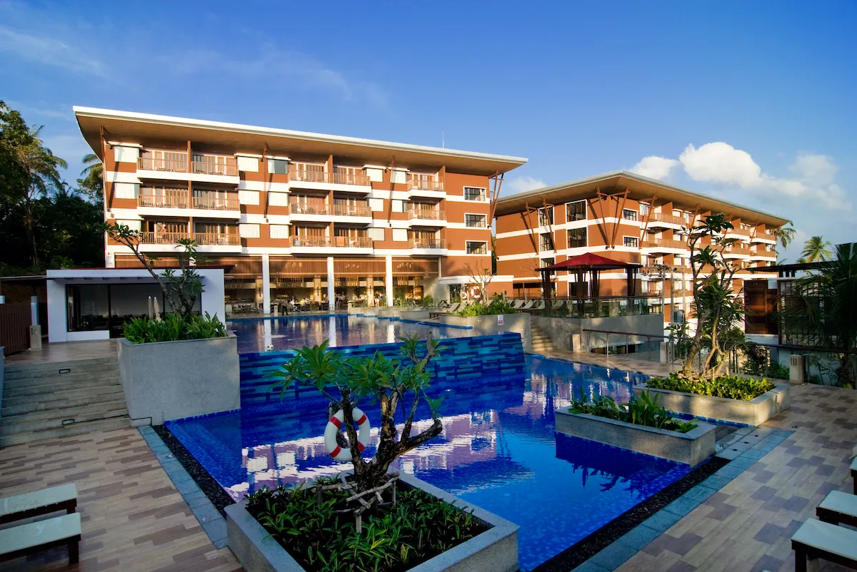 Scenic view of Peach Blossom Resort Phuket featuring a large outdoor swimming pool, landscaped sun deck and modern accommodation buildings under a clear blue sky