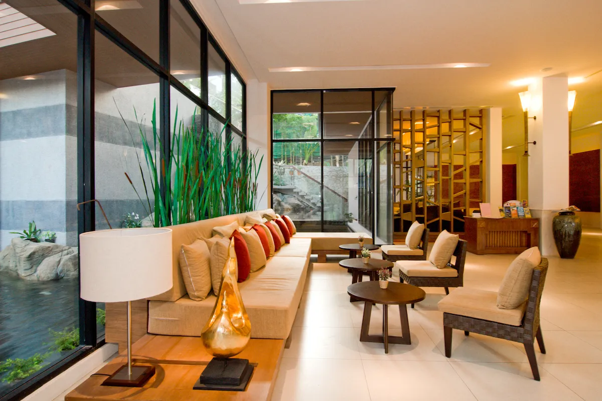 Stylish lobby lounge at Peach Blossom Resort Phuket featuring a long cushioned sofa with colourful pillows, modern decor and large glass windows overlooking a serene water feature