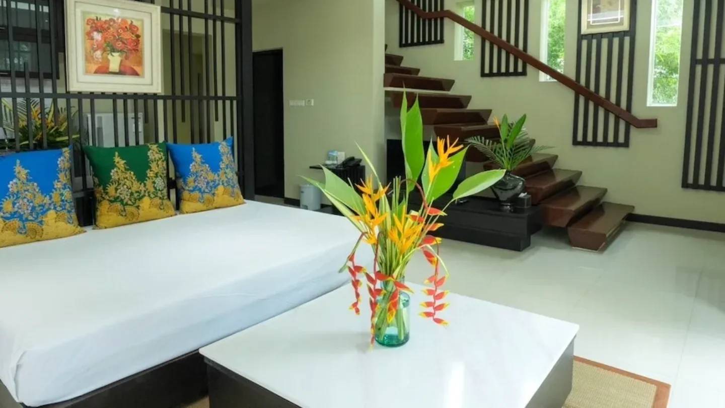 Stylish villa interior with a large bed, cushions, glass coffee table with tropical flowers, and a wooden staircase leading to the upper level