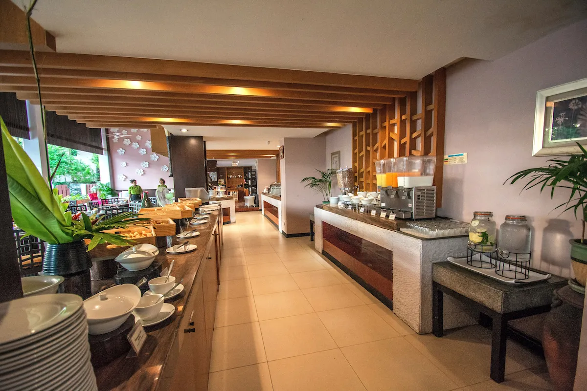 Bright and spacious dining area at Peach Blossom Resort featuring contemporary wooden decor, neatly arranged tables with orange cushioned chairs, warm ambient lighting, and chefs preparing food at the buffet counter in the background